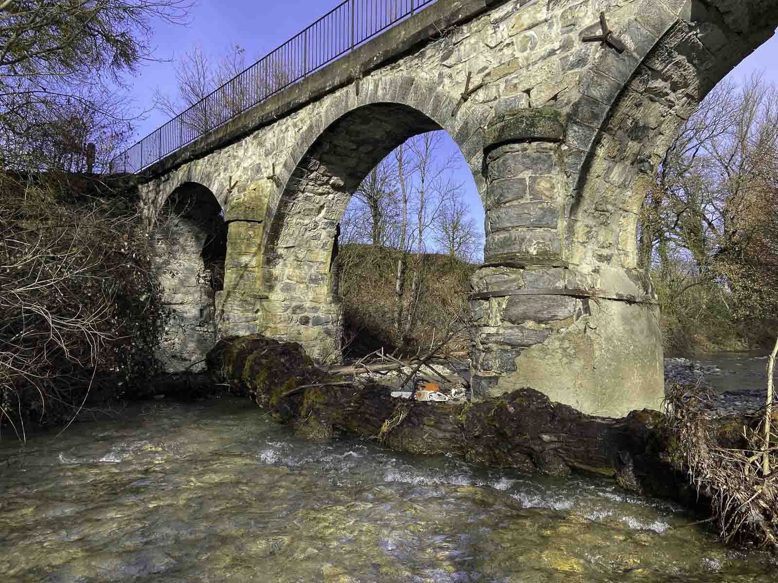 photo de l'aqueduc du Céans et du peuplier coincé contre ses piles avec en premier le lit de la rivière puis un gros tronc de peuplier en partie dans l'eau appuyé contre deux piles de l'aqueduc, visible au dessus du peuplier et en arrière plan quelques branches des arbres qui bordent la rivière en aval et le ciel bleu